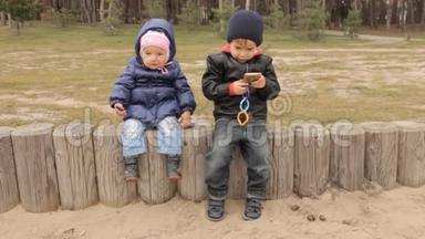 <strong>一个</strong>小男孩和<strong>一个</strong>小女孩坐在篱笆上，男孩<strong>按下</strong>智能手机上的按钮。 女孩摆动着腿。